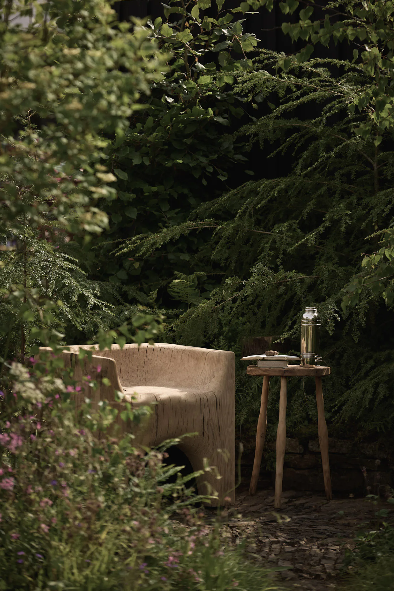 Carved timber chair and three-legged side table set among conifers and woodland planting